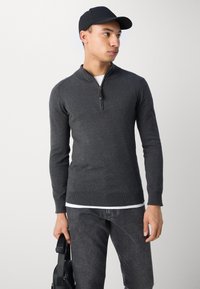 Tamno sivi zip-up pulover s rebrastim manžetama i rubom. Model nosi bijelu majicu ispod i drži crnu torbu. Jednostavan, prilagođen dizajn.