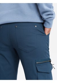 Niebieskie spodnie cargo z zamkniętą kieszenią i dwiema tylnymi kieszeniami. Materiał wydaje się gładki z delikatnym połyskiem. Szwy są widoczne wzdłuż kieszeni.