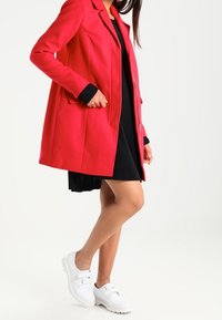 Abrigo rojo de manga larga con un diseño entallado, dos bolsillos de parche y cuello con muesca, combinado con un vestido negro plisado y zapatillas blancas.