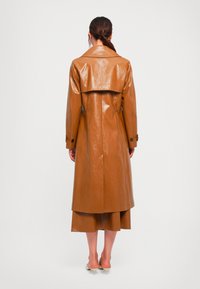 Donna che indossa un lungo trench di pelle marrone e una gonna coordinata, in piedi di spalle, con scarpe con tacco a kitten bianche su uno sfondo neutro.