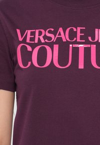 Violetinės medvilnės marškinėliai su dideliu, blizgiu rožiniu tekstu, skaitančiu "VERSACE JEAN COUTURE". Klasikinis apvalus kaklas ir trumpi rankoviai.