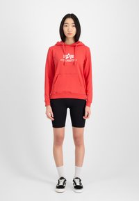 Sudadera roja con logo blanco, presenta un bolsillo canguro y capucha con cordón. Se lleva con pantalones cortos de ciclismo negros y zapatillas negras.
