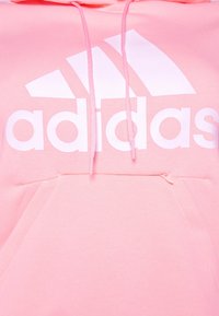 Felpa rosa in cotone con grande logo bianco Adidas, con cordini e una piccola tasca frontale con zip. Tessuto morbido, design informale.