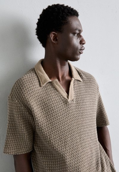 Camisa de polo beige texturizada con mangas cortas, cuello suave en contraste y un patrón de diamantes. El material parece tejido y casual.