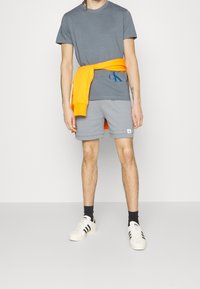 Grå bomullströja med blå logga, orange sweatshirt knuten i midjan, grå shorts med vit etikett och vita sneakers med svarta ränder.