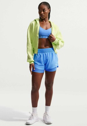 Kvinde står iført gul cropped hættetrøje, blå sports-bh, blå shorts, hvide sokker og hvide sneakers mod en ensfarvet baggrund.
