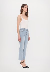 Top blanco sin mangas, jeans de tiro alto azul claro y tacones negros puntiagudos llevados por una modelo de pie de lado. Diseño minimalista, forma ajustada.