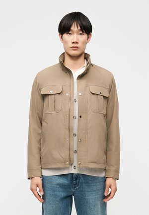 Junger Mann trägt eine beige Jacke mit Klappen-Brusttaschen über einem weißen, geknöpften Pullover und blauen Jeans, steht vor einem weißen Hintergrund.