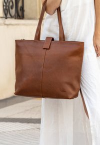 Une personne en robe blanche tenant un sac fourre-tout en cuir marron avec deux anses et une fermeture centrale à rabat, debout en plein air sur un sol pavé.