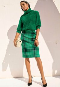 Groene fuzzy trui met cropped mouwen, gecombineerd met een groene geruite kokerrok. Zwarte hoge hakken en een geweven clutch maken de look compleet.