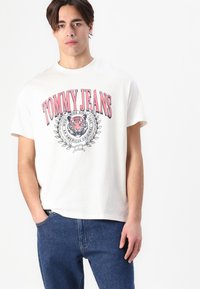 Cremefarvet bomulds T-shirt med fed rød 'TOMMY JEANS' logo, et tiger grafik og cirkulært blomster insignie. Bæret med blå jeans.