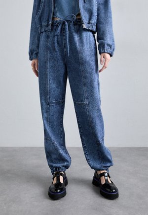 Donkerblauwe denim jogger pants met een elastische tailleband en een trekkoord. Kenmerken zijn contrasterende stiksels en elastische manchetten; gecombineerd met zwarte chunky schoenen.