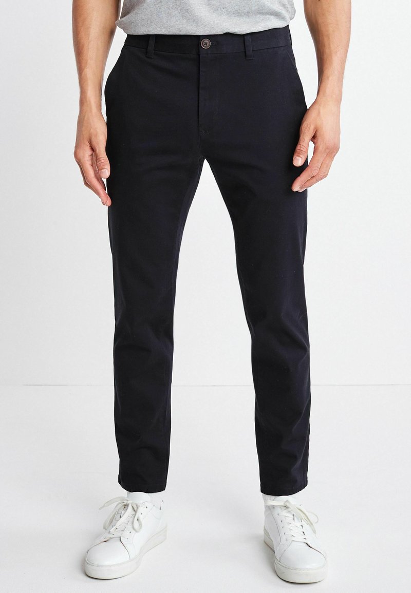 Marineblaue Baumwollchinos mit schmaler Passform, flachem Vorderteil und Knopfverschluss, kombiniert mit weißen Turnschuhen. Glatte Textur und minimales Design.