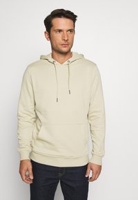 Beige hettegenser med en frontlomme, justerbare snorer, ribbede mansjetter og hem, laget av myk, teksturert stoff.