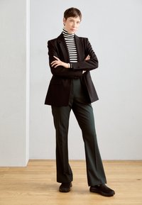 Blazer nero con un taglio sartoriale, dolcevita a righe, pantaloni verde scuro e scarpe slip-on nere. Tessuto liscio e linee pulite.
