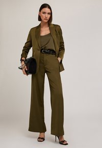 Costum satinat de culoare verde olive, cu un sacou ajustat, pantaloni evazați și un top asortat. Curea și geantă de mână negre cu detalii din lanț, completate cu pantofi cu toc negri.