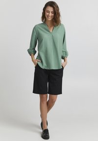 Lys grøn bluse med V-udskæring og 3/4 pufærmer parret med sorte knælange shorts. Outfittet har afslappede og lette elementer.
