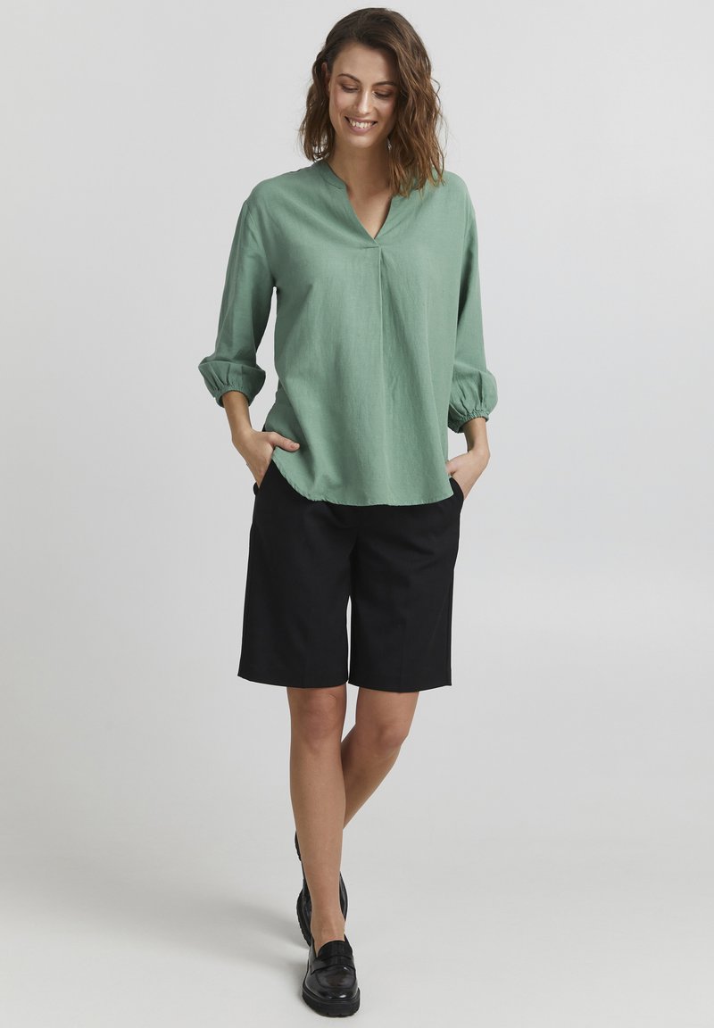 Lys grøn bluse med V-udskæring og 3/4 pufærmer parret med sorte knælange shorts. Outfittet har afslappede og lette elementer.