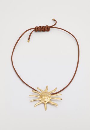 Colgante de color dorado en forma de sol con rasgos faciales, atado a un cordón trenzado marrón con nudos ajustables.
