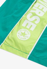 Τζάκετ αδιάβροχο σε τιρκουάζ και λαχανί πράσινο, με μεγάλο λευκό κείμενο "CONVERSE" και κυκλικό λογότυπο με το όνομα της μάρκας και το έμβλημα αστεριού.