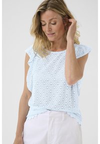 Top in cotone azzurro chiaro con design in broderie anglaise, bordi ondulati e maniche a farfalla, su uno sfondo neutro.