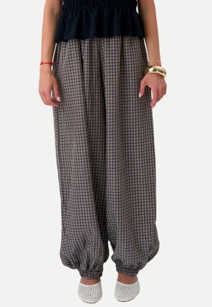 Femme portant un pantalon large à carreaux avec des poignets élastiques, un top noir, des chaussures blanches sans lacets et des bracelets aux deux poignets.
