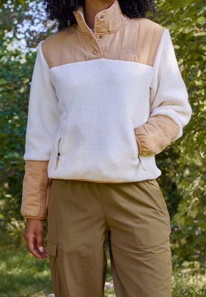 Vrouw draagt een lichtbruine en witte fleece trui met ritssluitingen in de zakken en lichtbruine cargobroek, staand buiten tussen groene bomen.