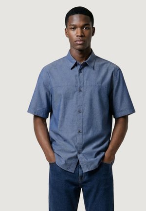 Młody mężczyzna stojący z rękami w kieszeniach, ubrany w niebieską koszulę na krótki rękaw z guzikami i ciemnoniebieskie dżinsy na jednolitym tle.