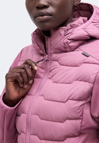 Personne portant une veste matelassée rose, tenant la tirette de la fermeture éclair près du col, se concentrant sur le torse supérieur et les détails de la main.