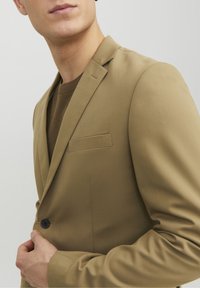 Beige kavaj med glipor, enkel knappstängning och ficka på bröstet. Slät tygtextur, figursydd design. Buren över en brun tröja.