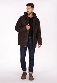 Brun hættejakke med lynlåse, flere lommer, båret over en sort skjorte. Parret med mørke jeans og brune snørebåndsstøvler.