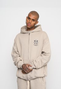 Felpa beige con zip, dotata di un ampio cappuccio, tasche frontali e un logo grafico sul petto. Il tessuto sembra morbido ed ha una vestibilità comoda.