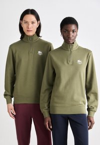 Kelių žalia ketvirčio užtrauktuko megztinis su nertomis rankogaliais ir apačia. Balta Lacoste logotipas kairėje krūtinės pusėje. Modelis derina su tamsiomis kelnėmis.