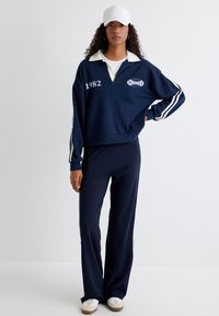 Donkerblauwe sweatshirt met witte kraag en streepdetails, met "1982" op de voorkant. Gecombineerd met donkere broeken en witte sneakers.