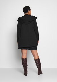 Melna mētelis ar plašu kapuci, plūstošs dizains, līdz viduklim garums, apvienots ar melnu rakstainu svārku un brūnām zābaciņām līdz ceļiem.