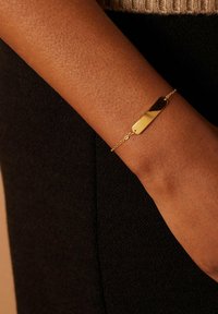 Goudkleurige armband met een rechthoekige hanger, featuring een gladde oppervlakte en een delicate ketting, gedragen op een gestructureerde huidkleur.