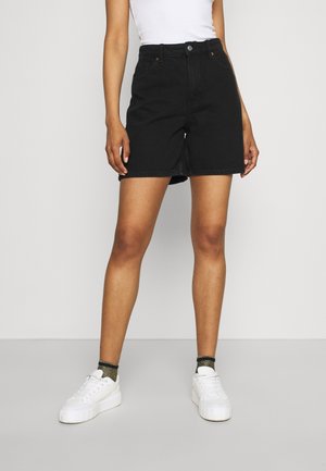 Persona con shorts de mezclilla negros, camiseta de tirantes blanca, calcetines negros con brillo y zapatillas blancas de plataforma, de pie contra un fondo claro y sencillo.