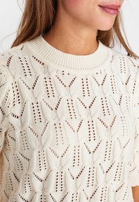 Beige stickad tröja med en strukturerad, öppna mönster och rund halsringning med korta ärmar. Designen inkluderar förlängda motiv.