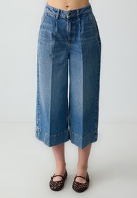 Pantalons culottes en denim bleu clair avec une taille haute, des jambes larges et des ourlets bruts, associés à des chaussures plates tissées noires.