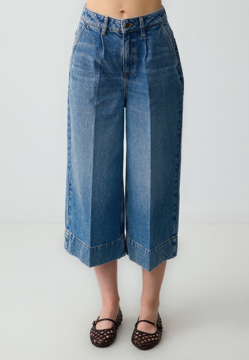 Pantalons culottes en denim bleu clair avec une taille haute, des jambes larges et des ourlets bruts, associés à des chaussures plates tissées noires.