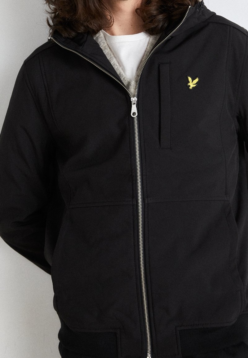 Zwarte jas met ritssluiting en capuchon, voorzien van een voorvak, geribbelde manchetten en een gele adelaar-logo op de borst. Glad, mat oppervlak.