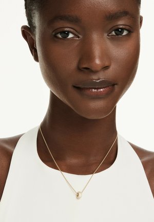 Collar de oro con un colgante circular texturizado, usado con una blusa sin mangas blanca. Presenta una cadena delicada y acentos brillantes.