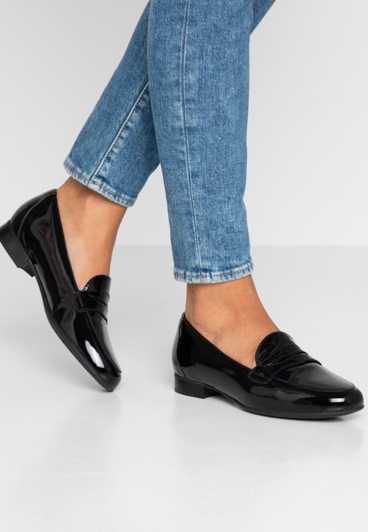 Mocassins en cuir verni noir portés avec un jean en denim bleu longueur cheville sur fond blanc.