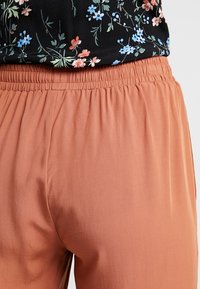 Persikofärgade lätta byxor med elastisk midja, som har en mjuk textur och en avslappnad passform. Blommönstrad topp delvis synlig.