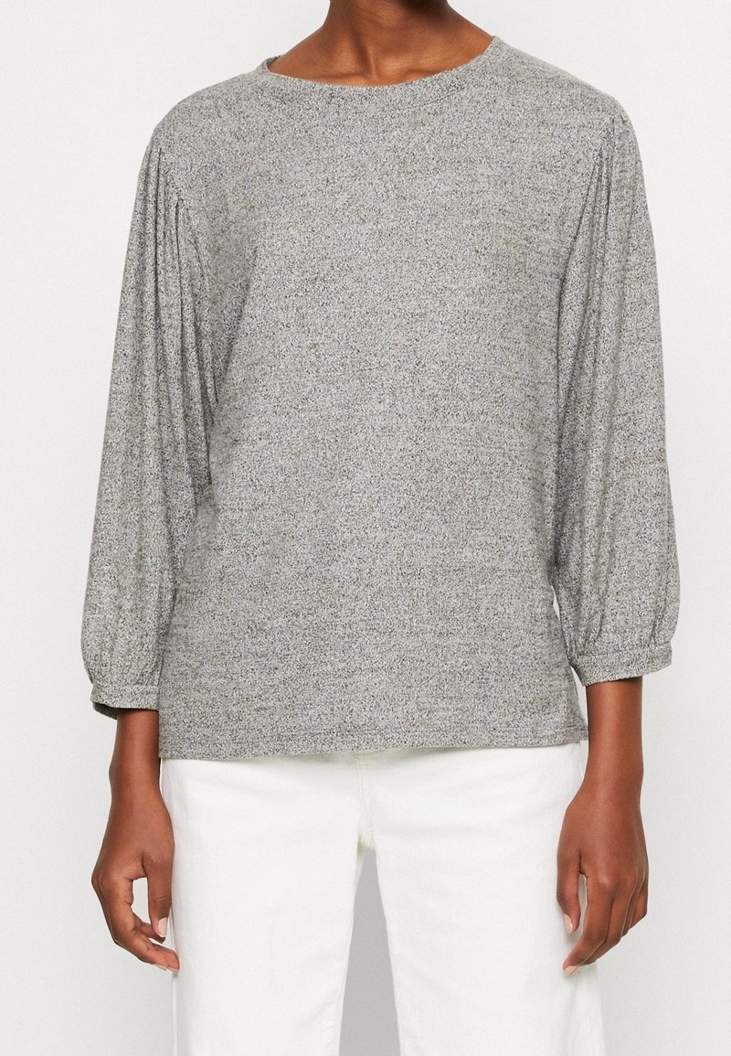 Maglione grigio con maniche lunghe e arricciate e scollatura tonda. Il tessuto presenta un sottile motivo a trama, abbinato a pantaloni bianchi aderenti.