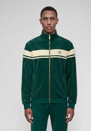 Homme portant une veste de survêtement en velours vert foncé avec des rayures horizontales crème et une fermeture éclair dorée, debout devant un fond clair uni.