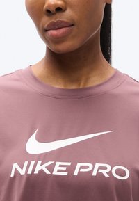 Tricou Nike Pro cu mâneci scurte, în roz prăfuit, având un logo mare alb și text. Materialul este neted, cu gât rotund și ușor lucios.