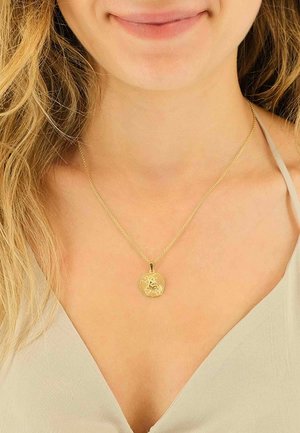 Collier en or avec un pendentif rond présentant un design détaillé. La chaîne est fine et délicate, reposant au-dessus d'un vêtement de couleur claire.