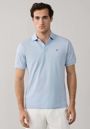 Jonge man staand, gekleed in een lichtblauw poloshirt met een klein donkerblauw logo van gekruiste peddels, en een witte broek, neutrale achtergrond.