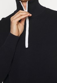 Czarny sweter z prążkowanej dzianiny z wysokim kołnierzem i białym detalem zamka błyskawicznego. Częściowo widoczna dłoń, podkreślająca strukturę tkaniny i dopasowanie.